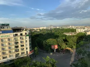 Bức ảnh chụp toàn cảnh một góc thành phố từ trên cao, có thể là thành phố Thanh Hóa, vào một buổi sáng trong lành. Khung cảnh bao gồm nhiều tòa nhà, đường phố và cây xanh tươi tốt. Ở phía xa là những dãy núi đá vôi đặc trưng của khu vực.。Các dòng chữ có thể nhận dạng được bao gồm:。- Trên tòa nhà cao tầng bên trái: biển hiệu lớn màu xanh lá cây "HOTEL", biển hiệu "NGÂN HÀNG Á CHÂU", và dòng chữ "Welcome to Palm Hotel" ở tầng trệt. Một biển hiệu khác màu đỏ ở góc tòa nhà có chữ "NHÀ SÁCH".。- Trên nóc một tòa nhà khác có chữ "THANH... GRO...".。- Một biển quảng cáo lớn màu đỏ của hãng bảo hiểm "GENERALI" với khẩu hiệu "BẢO HIỂM VÌ TIM Ý" và hashtag "#SốngNhưÝ".。- Một cổng chào màu đỏ bắc qua đường có các dòng chữ như "PHƯỜNG ĐIỆN BIÊN", "PHỐ LÊ HOÀN", "PHƯỜNG LAM SƠN".。- Một tòa nhà ở xa có biển hiệu "DAI VIET PALACE".。Hoạt động chính trong ảnh là giao thông thưa thớt trên đường phố, với một vài chiếc xe máy đang di chuyển. Cảnh vật cho thấy một khu đô thị có nhiều cây cối và không khí yên tĩnh.。（日本語訳）。上空から撮影された、爽やかな朝の都市の一角の全景写真で、おそらくタインホア市だと思われます。風景には多くの建物、道路、そして青々とした緑が含まれています。遠方には、この地域特有の石灰岩の山々が見えます。認識できる文字は以下の通りです：。- 左側の高層ビルには、大きな緑色の「HOTEL」の看板、「NGÂN HÀNG Á CHÂU」の看板、そして1階部分に「Welcome to Palm Hotel」の文字があります。建物の角にある別の赤い看板には「NHÀ SÁCH」という文字があります。- 別の建物の屋上には「THANH... GRO...」という文字があります。- 保険会社「GENERALI」の大きな赤い広告看板には、スローガン「BẢO HIỂM VÌ TIM Ý」とハッシュタグ「#SốngNhưÝ」が書かれています。- 道路をまたぐ赤い歓迎ゲートには、「PHƯỜNG ĐIỆN BIÊN」、「PHỐ LÊ HOÀN」、「PHƯỜNG LAM SƠN」といった文字があります。- 遠くの建物には「DAI VIET PALACE」の看板があります。写真の中の主な動きは、道路上のまばらな交通で、数台のバイクが移動しています。景色は、多くの木々があり静かな雰囲気の都市部であることを示しています。