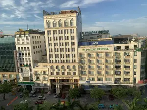 Bức ảnh chụp một góc phố sầm uất tại một trung tâm đô thị, nhìn từ trên cao xuống. Khung cảnh có nhiều tòa nhà cao tầng với các biển hiệu thương mại. Các văn bản có thể nhận dạng được bao gồm:。- Tên khách sạn: 'DAI VIET HOTEL', 'PALM HOTEL'.。- Tên spa: 'HOA SEN SPA'.。- Tên các ngân hàng: 'ACB' (NGÂN HÀNG Á CHÂU), 'OCB' (Ngân Hàng Phương Đông, CHUYỂN TIỀN QUỐC TẾ), 'SeABank'.。- Các biển hiệu khác: 'NHÀ SÁCH', 'MASSAGE' (biển hiệu dọc màu đỏ), 'Welcome to Palm Hotel'.。Hoạt động chính diễn ra ở dưới mặt đất là sinh hoạt đô thị. Có nhiều xe ô tô đang đỗ trong bãi và ven đường. Một vài người đang đứng cạnh những chiếc xe ô tô màu xanh ở góc dưới bên phải. Cây xanh được trồng dọc theo đường phố. Phía xa, có thể nhìn thấy hình dáng của những ngọn núi dưới bầu trời xanh có mây. Có một băng rôn màu đỏ treo ngang qua đường nhưng chữ viết trên đó không thể đọc được.。（日本語訳）。ある都市の中心部にある賑やかな街角を上から見下ろして撮影した写真です。風景には、商業看板のある高層ビルが多数あります。識別可能なテキストには以下が含まれます：。- ホテル名：「DAI VIET HOTEL」、「PALM HOTEL」。- スパ名：「HOA SEN SPA」。- 銀行名：「ACB」（アジア商業銀行）、「OCB」（東方銀行、国際送金）、「SeABank」。- その他の看板：「NHÀ SÁCH」（書店）、「MASSAGE」（赤い縦長の看板）、「Welcome to Palm Hotel」。地上で行われている主な活動は、都市の営みです。駐車場や道端には多くの自動車が駐車しています。右下隅では、数人が緑色の自動車の隣に立っています。通りに沿って緑の木々が植えられています。遠くには、雲のある青空の下に山々の姿が見えます。通りを横切って赤い横断幕が掲げられていますが、そこに書かれた文字は読み取れません。