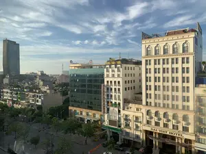 Đây là một bức ảnh chụp quang cảnh một khu đô thị sầm uất từ trên cao, có thể là thành phố Thanh Hóa. Bầu trời trong xanh với những gợn mây trắng. Có nhiều tòa nhà cao tầng với kiến trúc đa dạng. Ở phía trước bên phải là một khách sạn lớn màu trắng với dòng chữ "DAI VIET HOTEL". Bên cạnh đó là các tòa nhà khác có biển hiệu của các ngân hàng như "SeABank", "OCB" (Ngân Hàng Phương Đông). Một tòa nhà khác có biển hiệu màu vàng "HOA SEN SPA". Ở phía xa hơn một chút là một tòa nhà văn phòng bằng kính với logo và tên "VietinBank" trên nóc. Phía dưới là một con đường rộng rãi có cây xanh trồng hai bên, với một vài ô tô và xe máy đang lưu thông. Ở phía xa bên trái là một tòa nhà chọc trời và một tháp ăng-ten viễn thông.。（日本語訳）。これは、上空から撮影した賑やかな都市部の風景写真で、おそらくタインホア市だと思われます。空は青く澄み渡り、白い雲がいくつか浮かんでいます。多様な建築様式の高層ビルが数多くあります。右手前には「DAI VIET HOTEL」という文字が入った大きな白いホテルがあります。その隣には、「SeABank」や「OCB」（東方銀行）といった銀行の看板がある他のビルがあります。別のビルには「HOA SEN SPA」という黄色い看板があります。少し奥には、屋上に「VietinBank」のロゴと名前が入ったガラス張りのオフィスビルがあります。下には両側に街路樹が植えられた広い道路があり、数台の自動車やバイクが通行しています。左奥には、超高層ビルと通信アンテナ塔があります。