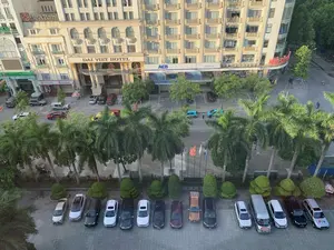 Bức ảnh được chụp từ góc nhìn trên cao, bao quát một khu phố đô thị tại Việt Nam. Cảnh quan chủ yếu là các tòa nhà cao tầng, đường phố và cây xanh. Có thể nhận dạng được nhiều văn bản trên các biển hiệu: tên khách sạn 'DAI VIET HOTEL' và một biển chào 'Welcome to Palm Hotel'. Nhiều chi nhánh ngân hàng cũng có mặt với các biển hiệu lớn như 'ACB' (NGÂN HÀNG Á CHÂU), 'SeABank', và 'OCB' (Ngân Hàng Phương Đông) có ghi 'CHUYỂN TIỀN QUỐC TẾ'. Một biển hiệu màu đỏ ở góc phố có chữ 'NHÀ...' nhưng phần còn lại bị che khuất. Về hoạt động, đường phố có xe ô tô và xe máy đang lưu thông. Nhiều ô tô đang đỗ trong các bãi đỗ xe được quy hoạch trước các tòa nhà. Một chiếc xe van có logo 'PTV'. Một hàng cây cọ cao chạy dọc theo vỉa hè, xen kẽ là các bụi cây được cắt tỉa. Phía trước có một dãy cột cờ, trong đó có lá cờ đỏ sao vàng của Việt Nam. Một vài người dân đang đi lại trên đường và vỉa hè.。（日本語訳）。写真は上からの視点で撮影されており、ベトナムのある都市の一角を捉えています。主な景観は高層ビル、道路、そして緑の木々です。看板には多くの文字が認識できます：ホテル名「DAI VIET HOTEL」と「Welcome to Palm Hotel」という歓迎の看板。多くの銀行支店も存在し、「ACB」（アジア商業銀行）、「SeABank」、そして「国際送金」と書かれた「OCB」（オリエント商業銀行）のような大きな看板があります。角にある赤い看板には「NHÀ...」という文字がありますが、残りの部分は隠れています。交通状況については、道路には自動車やバイクが通行しています。多くの自動車が建物の前に計画的に設けられた駐車場に停まっています。「PTV」のロゴが入ったバンが一台あります。高いヤシの木が歩道に沿って一列に並び、その間には刈り込まれた低木があります。前方には旗竿が並んでおり、その中にはベトナムの金星紅旗（黄色の星の入った赤旗）があります。何人かの人々が道路や歩道を行き来しています。
