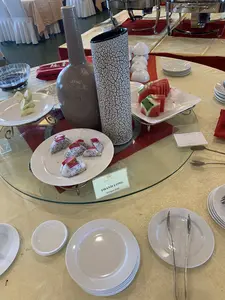 Bối cảnh là một quầy đồ ăn tự chọn (buffet) trong một nhà hàng hoặc phòng tiệc. Trên một chiếc bàn tròn trải khăn màu vàng, có một kệ kính trưng bày các món tráng miệng. Một đĩa sứ trắng đựng các miếng thanh long đã được cắt sẵn. Phía trước đĩa thanh long có một tấm biển ghi "SAO MAI", "THANH LONG", và "Dragon fruit". Ngoài ra còn có một đĩa đựng dưa hấu và một đĩa đựng ổi đã được cắt miếng. Xung quanh bàn có sắp xếp sẵn các chồng đĩa sứ trắng, bát nhỏ, kẹp gắp và dĩa để khách tự phục vụ. Ở phía sau, có thể thấy các dụng cụ hâm nóng thức ăn bằng kim loại và các bàn ăn khác được trải khăn, cùng những chiếc ghế được bọc vải trắng.。（日本語訳）。背景は、レストランまたは宴会場にあるビュッフェカウンターです。黄色いテーブルクロスが敷かれた円卓の上には、デザートを陳列するガラスの棚があります。白い磁器の皿に、カット済みのドラゴンフルーツの切り身が盛られています。ドラゴンフルーツの皿の前には、「SAO MAI」、「THANH LONG」、「Dragon fruit」と書かれたプレートがあります。その他にも、スイカを盛った皿と、カットされたグアバを盛った皿があります。テーブルの周りには、客がセルフサービスで取れるように、積み重ねられた白い磁器の皿、小鉢、トング、フォークが用意されています。後方には、金属製のフードウォーマーや、テーブルクロスが敷かれた他の食卓、そして白い布で覆われた椅子が見えます。