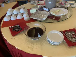 Bối cảnh là một bàn tiệc buffet trong nhà, có thể là ở một nhà hàng hoặc một sự kiện. Bàn ăn hình tròn được phủ khăn trải bàn màu vàng và váy bàn màu đỏ. Trên bàn có bày biện sẵn các món tráng miệng và dụng cụ ăn uống. Có nhiều chồng bát và đĩa sứ màu trắng. Thìa và dĩa được xếp gọn gàng trên khăn ăn màu đỏ. Ở trung tâm bàn, có một tô thủy tinh lớn đựng một món chè màu sẫm, bên cạnh có một tấm biển nhỏ ghi 'SAO MAI' và 'CHÈ ĐẬU ĐEN' (black bean sweet soup). Ngoài ra, trên các kệ tầng bằng kính còn có các đĩa trái cây đã được cắt sẵn như thanh long, ổi, và dưa hấu. Một khay thức ăn được đậy bằng giấy bạc. Toàn bộ khung cảnh cho thấy một bữa tiệc tự chọn đã được chuẩn bị để phục vụ khách.。（日本語訳）。背景は屋内のビュッフェテーブルで、レストランかイベント会場のようです。円形のテーブルには黄色のテーブルクロスと赤色のテーブルスカートがかけられています。テーブルの上にはデザートと食器が用意されています。白い磁器のボウルと皿がたくさん重ねられています。スプーンとフォークは赤いナプキンの上にきれいに並べられています。テーブルの中央には、濃い色のチェーが入った大きなガラスのボウルがあり、その隣には「SAO MAI」と「CHÈ ĐẬU ĐEN」（黒豆のチェー）と書かれた小さな札があります。その他に、ガラスの段になった棚には、ドラゴンフルーツ、グアバ、スイカなど、カット済みの果物の皿があります。アルミホイルで覆われた料理のトレイが一つあります。全体の光景は、客をもてなすために準備されたビュッフェパーティーを示しています。