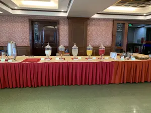 Bối cảnh là một khu vực phục vụ đồ ăn uống tự chọn, có thể là trong một khách sạn hoặc trung tâm hội nghị. Một chiếc bàn dài được phủ khăn trải bàn màu đỏ, trên đó bày biện sẵn sàng một bữa tiệc trà hoặc bữa sáng.。Trên bàn có nhiều loại đồ uống và thức ăn nhẹ. Từ trái sang phải, có một bình giữ nhiệt lớn bằng inox, tiếp theo là năm bình thủy tinh lớn có vòi chứa các loại nước giải khát. Các loại nước này có màu sắc khác nhau: trắng đục, nâu nhạt, trong suốt, vàng cam và đỏ. Phía trước mỗi bình có các tấm biển nhỏ ghi tên đồ uống, một số có thể đọc được là "NƯỚC ĐẬU NÀNH", "TRÀ ĐÁ", "NƯỚC CAM", "NƯỚC DƯA HẤU".。Ở phía cuối bàn bên phải, có một máy nướng bánh mì sandwich màu trắng, hai bình thủy tinh đựng trà hoặc cà phê, một giỏ đựng bánh mì sandwich đã cắt lát, cùng với bơ và mứt. Phía sau bàn tiệc là bức tường dán giấy hoa văn màu hồng và các chi tiết trang trí bằng gỗ. Có một cánh cửa gỗ lớn và một lối đi khác có biển báo "EXIT" màu xanh lá cây phát sáng.。（日本語訳）。背景は、ホテルまたは会議センター内にあると思われる、セルフサービスの飲食エリアです。赤いテーブルクロスで覆われた長いテーブルの上に、ティーパーティーまたは朝食が準備されています。テーブルの上には、多くの種類の飲み物と軽食があります。左から右へ、ステンレス製の大きな保温ポットが一つあり、続いて様々な種類のソフトドリンクが入った蛇口付きの大きなガラス製の瓶が5つあります。これらの飲み物は、乳白色、薄茶色、透明、オレンジがかった黄色、赤色と、それぞれ異なる色をしています。それぞれの瓶の前には飲み物の名前が書かれた小さな札があり、そのうちいくつかは「NƯỚC ĐẬU NÀNH」（豆乳）、「TRÀ ĐÁ」（アイスティー）、「NƯỚC CAM」（オレンジジュース）、「NƯỚC DƯA HẤU」（スイカジュース）と読むことができます。テーブルの右端には、白いサンドイッチトースターが一つ、お茶またはコーヒーが入ったガラス製のポットが二つ、スライスされたサンドイッチ用のパンが入った籠が一つ、そしてバターとジャムがあります。ビュッフェテーブルの後ろには、ピンク色の模様の壁紙が貼られた壁と、木製の装飾があります。大きな木製のドアが一つと、光る緑色の「EXIT」の標識がある別の通路があります。