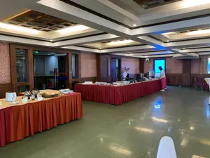 Bối cảnh là một phòng ăn tự chọn, có thể là của một khách sạn, đang phục vụ bữa sáng. Có hai dãy bàn dài được phủ khăn trải bàn màu đỏ và cam, bày biện đồ ăn theo kiểu buffet. Trên dãy bàn chính, một nhân viên mặc đồng phục đầu bếp đang đứng sau quầy, nơi có các nồi và khay hâm nóng thức ăn đang bốc hơi. Một vị khách nữ mặc áo phông trắng và quần hồng đang lấy thức ăn vào bát. Nhiều chồng đĩa và bát sứ trắng được xếp sẵn cho khách. Một bàn nhỏ hơn ở phía trước có máy nướng bánh mì, các bình thủy tinh đựng trà hoặc cà phê, một giỏ bánh mì sandwich, cùng với bơ và mứt. Không gian phòng ăn có sàn lát gạch bóng, tường dán giấy hoa văn và ốp gỗ, trần nhà được thiết kế giật cấp với hệ thống đèn chiếu sáng. Về chữ viết, có một biển báo "EXIT" màu xanh lá cây phía trên cửa ra vào. Trên cửa kính có dòng chữ "LƎTOH IAM OAS", có khả năng là tên "SAO MAI HOTEL" nhìn ngược từ bên trong.。（日本語訳）。背景は、ホテルのものと思われるセルフサービスのダイニングルームで、朝食を提供しています。赤とオレンジのテーブルクロスがかけられた長いテーブルが2列あり、ビュッフェ形式で料理が並べられています。メインのテーブル列では、シェフの制服を着たスタッフがカウンターの後ろに立っており、そこには湯気の立つ鍋や料理の保温トレイがあります。白いTシャツとピンクのズボンを履いた女性客が、ボウルに食べ物を取っています。たくさんの白い磁器の皿とボウルが、客のために積み重ねられています。手前にある小さなテーブルには、トースター、お茶またはコーヒーが入ったガラスポット、サンドイッチパンのバスケット、そしてバターとジャムがあります。ダイニングルームの空間は、光沢のあるタイル張りの床、花柄の壁紙と木製パネルの壁、照明システムを備えた段差のあるデザインの天井が特徴です。文字については、出入り口の上に緑色の「EXIT」の看板があります。ガラスのドアには「LƎTOH IAM OAS」という文字があり、これは内側から逆さに見た「SAO MAI HOTEL」という名前である可能性があります。