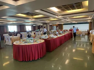 Bối cảnh là một phòng ăn rộng rãi, có thể là của một nhà hàng hoặc khách sạn, nơi đang diễn ra một bữa tiệc buffet. Một dãy bàn dài được phủ khăn trải bàn màu đỏ, trên đó bày rất nhiều nồi hâm nóng thức ăn (chafing dish) bằng inox sáng bóng, cùng với các chồng đĩa và bát sứ trắng. Mọi người đang đứng lựa chọn và lấy thức ăn từ quầy buffet. Ở phía trước, có một bàn tròn riêng bày các món khai vị như salad và thịt nguội; trên bàn này có một tấm biển nhỏ ghi chữ 'SALAD RAU TRỘN'. Xung quanh phòng là nhiều bộ bàn ghế ăn uống khác, các bàn được phủ khăn màu vàng và ghế được bọc vải trắng. Sàn nhà lát gạch bóng loáng và trần nhà có thiết kế giật cấp với hệ thống đèn âm trần.。（日本語訳）。背景は、レストランかホテルのものと思われる広々としたダイニングルームで、ビュッフェ形式のパーティーが開催されています。赤いテーブルクロスで覆われた長いテーブルが並んでおり、その上には光沢のあるステンレス製のチェーフィングディッシュ（料理保温鍋）がたくさん並べられ、白い磁器の皿やボウルが積み重ねられています。人々はビュッフェカウンターから立って料理を選び、取っています。手前には、サラダやハムなどの前菜が並べられた別の円卓があり、このテーブルには「SALAD RAU TRỘN」（ミックスサラダ）と書かれた小さな札があります。部屋の周りには、他のダイニングテーブルと椅子のセットがたくさんあり、テーブルは黄色のクロスで、椅子は白い布で覆われています。床は光沢のあるタイル張りで、天井はダウンライトシステムを備えた段差のあるデザインです。