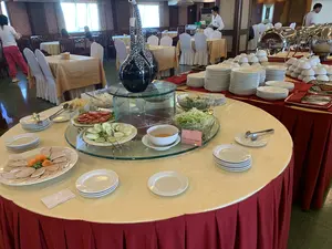 Bối cảnh là bên trong một nhà hàng hoặc phòng ăn của khách sạn, nơi đang phục vụ tiệc buffet. Ở tiền cảnh, một bàn tròn lớn được phủ khăn trải bàn màu vàng và váy bàn màu đỏ, bày biện các món ăn. Trên bàn có một đĩa chả lụa thái lát, một đĩa cà chua, một đĩa dưa chuột, và các bát salad. Một tấm biển nhỏ màu hồng ghi chữ 'SALAD RAU TRỘN' và 'Mixed salad'. Xung quanh các món ăn là các chồng đĩa sứ trắng sạch sẽ. Ở phía sau là dãy bàn buffet dài hơn, đặt các khay hâm nóng thức ăn bằng inox có nắp đậy, cùng với rất nhiều bát và đĩa được xếp chồng lên nhau. Trong không gian nhà hàng, có các nhân viên mặc đồng phục đang chuẩn bị và một vài thực khách đang ngồi ở các bàn ăn được sắp xếp sẵn.。（日本語訳）。背景は、ビュッフェが提供されているレストランまたはホテルのダイニングルームの中です。手前には、黄色のテーブルクロスと赤色のテーブルスカートで覆われた大きな円卓があり、料理が並べられています。テーブルの上には、スライスされたチャールアの皿、トマトの皿、きゅうりの皿、そしてサラダのボウルがあります。ピンク色の小さな札には「SALAD RAU TRỘN」と「Mixed salad」という文字が書かれています。料理の周りには、清潔な白い磁器の皿が積み重ねられています。後方には、より長いビュッフェテーブルの列があり、蓋付きのステンレス製フードウォーマーが置かれ、たくさんのボウルや皿が積み重ねられています。レストランの空間には、制服を着たスタッフが準備をしており、数人の客がセッティング済みのテーブルに座っています。