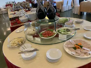 Trên các biển tên món ăn có ghi: "SAO MAI", "SALAD NGA", "Russian salad"; "SAO MAI", "SALAD CÀ CHUA", "Cherry tomatoes salad"; và "SAO MAI", "SALAD DƯA CHUỘT", "Cucumber salad".。Bối cảnh là một phòng ăn hoặc phòng tiệc trong nhà, có thể là của một nhà hàng hoặc khách sạn, nơi đang tổ chức một bữa tiệc buffet. Trên một chiếc bàn tròn và một dãy bàn dài được phủ khăn trải bàn màu vàng, có rất nhiều món ăn được bày ra. Các món ăn có thể nhận thấy bao gồm một đĩa giò lụa thái lát, salad dưa chuột, salad cà chua, salad Nga, và một đĩa bánh màu vàng. Đĩa và bát sứ trắng được xếp thành chồng, cùng với dao, dĩa, thìa để khách tự lấy. Ở phía sau, có các khay hâm nóng thức ăn bằng inox và một vài người đang đứng gần đó, dường như đang tham dự bữa tiệc.。（日本語訳）。料理名の札には、「SAO MAI」、「SALAD NGA」、「Russian salad」；「SAO MAI」、「SALAD CÀ CHUA」、「Cherry tomatoes salad」；そして「SAO MAI」、「SALAD DƯA CHUỘT」、「Cucumber salad」と書かれています。背景は、屋内のダイニングルームまたは宴会場で、レストランかホテルのものでしょうか、ビュッフェパーティーが開催されています。黄色いテーブルクロスで覆われた円卓と長いテーブルの上には、たくさんの料理が並べられています。見て取れる料理には、スライスされたベトナム風ポークソーセージ（ジョールア）の皿、きゅうりサラダ、トマトサラダ、ロシアサラダ、そして黄色いケーキの皿が含まれます。白い陶器の皿とボウルが重ねて置かれ、客が自分で取れるようにナイフ、フォーク、スプーンが添えられています。後方には、ステンレス製のフードウォーマーがあり、その近くに数人が立っており、パーティーに参加しているようです。