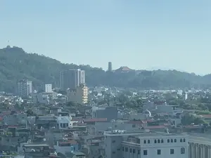 Đây là một bức ảnh chụp toàn cảnh một khu vực đô thị hoặc thành phố, nhìn từ một vị trí trên cao. Phía trước là một khu dân cư dày đặc với nhiều nhà cửa san sát nhau. Ở phía sau là một ngọn đồi lớn được bao phủ bởi cây xanh. Nổi bật trên đỉnh đồi là một công trình kiến trúc tôn giáo, bao gồm một ngôi chùa có mái ngói đỏ và một ngọn tháp nhiều tầng. Xen kẽ trong thành phố là một số tòa nhà cao tầng. Bầu trời trong ảnh có màu xanh nhạt và hơi mờ. Không có chữ viết nào có thể đọc được và không quan sát thấy hoạt động cụ thể nào của con người do góc chụp từ xa.。（日本語訳）。これは、ある都市部または都市の全景を高い場所から撮影した写真です。手前には、多くの家が密集している住宅地があります。後方には、緑に覆われた大きな丘があります。丘の頂上には、赤い瓦屋根の寺院と多層の塔を含む、宗教的な建築物が際立っています。都市の中には、いくつかの高層ビルが点在しています。写真の空は薄い青色で、少し霞んでいます。読み取れる文字はなく、遠くからの撮影のため、特定の人の活動は観察されません。