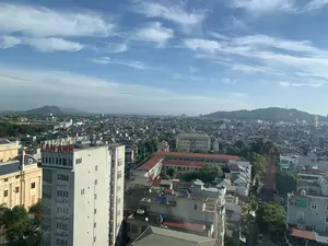 Bức ảnh chụp toàn cảnh một khu vực đô thị, có thể là thành phố Thanh Hóa, nhìn từ trên cao vào một ngày trời quang đãng. Bầu trời trong xanh với những gợn mây trắng. Quang cảnh bao gồm rất nhiều tòa nhà và nhà ở san sát nhau, cho thấy mật độ dân cư cao. Các công trình có kiến trúc đa dạng, từ nhà ống hiện đại đến các tòa nhà lớn hơn. Nhiều mái nhà có màu ngói đỏ, trong khi những mái nhà khác là mái bằng được trang bị các bình nước nóng năng lượng mặt trời và cục nóng điều hòa. Cây xanh được trồng xen kẽ giữa các công trình. Ở phía xa, có thể thấy những ngọn núi và đồi, một trong số đó có một tháp truyền thông trên đỉnh. Ở phía trước bên trái, có một tòa nhà cao tầng màu trắng, trên nóc có một biển hiệu lớn màu đỏ với chữ "LAN ANH". Không có hoạt động cụ thể nào của con người được ghi lại rõ ràng, chỉ là một khung cảnh tĩnh của thành phố.。（日本語訳）。晴れた日に上空から撮影された、おそらくタインホア市と思われる都市部の全景写真です。空は青く澄み渡り、白い雲がいくつか浮かんでいます。風景には多くの建物や住宅が密集しており、人口密度の高さがうかがえます。建物は、現代的なチューブハウスからより大きなビルまで、多様な建築様式が見られます。多くの屋根は赤い瓦葺きですが、他の屋根は平らで、太陽熱温水器やエアコンの室外機が設置されています。建物の間には緑の木々が点在して植えられています。遠くには山や丘が見え、そのうちの一つの頂上には通信塔があります。左手前には白い高層ビルがあり、その屋上には「LAN ANH」と書かれた大きな赤い看板があります。特定の人間の活動ははっきりと記録されておらず、ただ都市の静的な風景が写っているだけです。