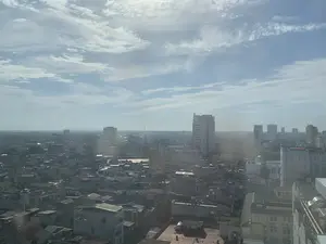 Đây là một bức ảnh chụp toàn cảnh một khu vực đô thị, có khả năng là thành phố Thanh Hóa, được chụp từ một vị trí trên cao. Khung cảnh cho thấy một sự kết hợp giữa các tòa nhà thấp tầng, nhà dân có mái ngói hoặc mái bằng, và các tòa nhà cao tầng hiện đại ở phía xa. Các công trình xây dựng nằm san sát nhau, tạo nên một quang cảnh đô thị dày đặc. Bầu trời trong xanh, có mây trắng và ánh nắng, tuy nhiên có một lớp sương mờ nhẹ. Trên nóc của nhiều tòa nhà có thể nhìn thấy các bồn chứa nước. Không có hoạt động cụ thể của con người hay biển hiệu, văn bản nào có thể nhận dạng được do khoảng cách chụp xa.。（日本語訳）。これは、ある都市部、おそらくタインホア市を、高い場所から撮影したパノラマ写真です。風景は、低層の建物、瓦屋根または陸屋根の民家、そして遠くにある近代的な高層ビルが混在している様子を示しています。建物は密集して建っており、高密度の都市景観を形成しています。空は青く澄み、白い雲と日差しがありますが、薄い霧がかかっています。多くの建物の屋上には貯水タンクが見えます。撮影距離が遠いため、特定の人間の活動や、識別できる看板や文字はありません。