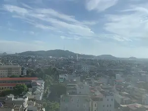 Bức ảnh chụp toàn cảnh một thành phố, có thể là thành phố Thanh Hóa, nhìn từ trên cao. Khung cảnh cho thấy một khu vực đô thị đông đúc với vô số nhà cửa và các tòa nhà san sát nhau. Các công trình có kiến trúc đa dạng, từ nhà ở thấp tầng với mái ngói hoặc mái tôn đến các tòa nhà cao tầng hơn. Xen kẽ giữa các khu dân cư là những hàng cây xanh. Ở phía xa là những ngọn đồi núi trập trùng được cây cối bao phủ. Trên đỉnh một ngọn đồi nổi bật có một công trình giống như tháp truyền hình. Bầu trời trong xanh, có nhiều mây trắng mỏng. Không có văn bản hay biển hiệu nào có thể đọc được và không thể xác định các hoạt động cụ thể của con người do góc nhìn bao quát và khoảng cách xa.。（日本語訳）。ある都市、おそらくタインホア市を上空から撮影したパノラマ写真。その光景は、無数の家屋や建物が密集する、人口の多い都市部を示している。建物は、瓦屋根やトタン屋根の低層住宅から、より高層のビルまで、多様な建築様式が見られる。住宅街の間には、緑の並木が点在している。目立つ丘の頂上には、テレビ塔のような建造物がある。空は青く澄んでおり、薄い白い雲が多く浮かんでいる。広角な視点と遠距離のため、読み取れる文字や看板はなく、人々の具体的な活動を特定することはできない。