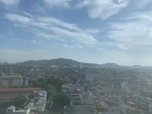 Đây là một bức ảnh chụp toàn cảnh một khu vực đô thị, có thể là thành phố Thanh Hóa, nhìn từ trên cao. Khung cảnh cho thấy một mật độ dày đặc các tòa nhà và nhà ở với nhiều loại mái khác nhau, nằm san sát nhau. Xen kẽ giữa các công trình xây dựng là những mảng cây xanh. Ở phía xa, có những ngọn đồi và núi được bao phủ bởi cây cối. Trên đỉnh một trong những ngọn đồi nổi bật có một công trình trông giống như tháp truyền hình hoặc tháp viễn thông. Bầu trời trong xanh với những đám mây trắng, mỏng và có một lớp sương mù nhẹ bao phủ thành phố. Không có chữ viết hay hoạt động cụ thể nào của con người có thể được nhận dạng trong ảnh.。（日本語訳）。これは、ある都市部、おそらくタインホア市を上空から撮影したパノラマ写真です。この風景は、様々な種類の屋根を持つ建物や家屋が密集している様子を示しています。建造物の間には、緑の木々が点在しています。遠方には、木々に覆われた丘や山があります。目立つ丘の一つには、テレビ塔か通信塔のような建造物があります。空は青く澄んでおり、薄い白雲があり、街は薄い霧に覆われています。