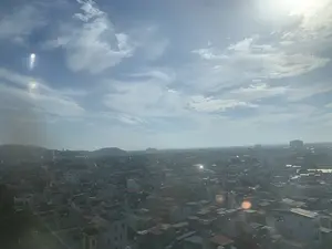 Bức ảnh chụp toàn cảnh một thành phố từ trên cao, có thể là thành phố Thanh Hóa. Khung cảnh cho thấy một khu vực đô thị dày đặc với vô số các tòa nhà và nhà ở san sát nhau, có mái nhà với nhiều kiểu dáng và màu sắc. Ở phía xa, có thể nhìn thấy những ngọn đồi hoặc núi thấp. Bầu trời trong xanh, có mây trắng và ánh nắng mặt trời chiếu sáng mạnh, tạo ra hiện tượng lóa ống kính. Do góc chụp từ xa nên không thể quan sát rõ các hoạt động cụ thể của con người. Không có văn bản hay biển hiệu nào có thể đọc được trong ảnh.。（日本語訳）。上空から撮影されたある都市の全景写真で、おそらくタインホア市だと思われます。その光景は、無数の建物や住宅が密集し、様々な形や色の屋根を持つ、高密度な都市部を示しています。遠方には、低い丘または山々が見えます。空は青く澄み、白い雲があり、太陽の光が強く差し込み、レンズフレア現象を引き起こしています。遠くからの撮影角度のため、人々の具体的な活動をはっきりと観察することはできません。写真の中には、読み取れる文字や看板はありません。