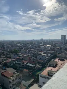 Đây là một bức ảnh chụp toàn cảnh một thành phố từ trên cao, có thể là thành phố Thanh Hóa. Khung cảnh cho thấy một khu vực đô thị đông đúc với vô số các tòa nhà và nhà ở san sát nhau. Các ngôi nhà có nhiều kiểu dáng và chiều cao khác nhau, với các loại mái đa dạng như mái ngói đỏ, mái tôn và mái bằng bê tông. Ở phía xa, có một vài tòa nhà cao tầng, trong đó có một tòa nhà ở bên phải có chữ viết trên đó nhưng không thể đọc rõ. Phía sau thành phố là những dãy núi mờ ảo. Bầu trời trong xanh, có mây trắng và ánh nắng mặt trời chiếu sáng rực rỡ. Không có hoạt động cụ thể của con người nào có thể được quan sát rõ ràng trong ảnh.。（日本語訳）。これは上空から撮影されたある都市の全景写真で、おそらくタインホア市だと思われます。この風景は、無数の建物や住宅が密集する、人口の多い都市部を示しています。家々は様々な様式と高さを持ち、赤い瓦屋根、トタン屋根、コンクリートの陸屋根など、多様な種類の屋根があります。空は青く澄み渡り、白い雲が浮かび、太陽の光が明るく輝いています。写真の中では、特定の人の活動をはっきりと観察することはできません。