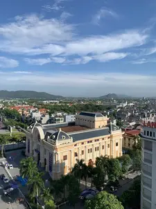 Bức ảnh chụp toàn cảnh một góc thành phố từ trên cao vào một ngày trời trong xanh. Đối tượng nổi bật ở trung tâm là một tòa nhà lớn, có kiến trúc cổ điển kiểu Pháp, sơn màu vàng nhạt với mái màu sẫm, đây là một nhà hát. Phía trước nhà hát có treo các băng rôn dọc màu tím, trên đó có thể đọc được dòng chữ: 'LIÊN HOAN TUỒNG VÀ DÂN CA KỊCH'. Xung quanh nhà hát là đường phố có một vài chiếc ô tô, nhiều cây xanh bao gồm cả cây cọ. Ở phía xa là quang cảnh thành phố với nhiều nhà dân và các tòa nhà khác, và ở đường chân trời là những dãy núi. Bên phải có một tòa nhà cao tầng với biển hiệu có chữ 'LAN' màu đỏ nhưng không nhìn rõ toàn bộ. Dưới đường có một vài người đang đi bộ và một số lều bạt được dựng tạm.。（日本語訳）。青空の日に、上空から街の一角を撮影した全景写真です。中央で際立っているのは、フランスの古典的な建築様式を持つ大きな建物で、薄い黄色に塗られ、屋根は濃い色をしています。これは劇場です。劇場の正面には紫色の縦長の横断幕が掲げられており、「LIÊN HOAN TUỒNG VÀ DÂN CA KỊCH」という文字が読み取れます。劇場の周りには、数台の車が走る道路や、ヤシの木を含む多くの緑の木々があります。遠方には、多くの民家や他の建物が立ち並ぶ街の景色が広がり、地平線には山々が連なっています。右側には高層ビルがあり、赤い文字で「LAN」と書かれた看板がありますが、全体ははっきりと見えません。路上には、数人の歩行者と、いくつか仮設のテントが設営されています。