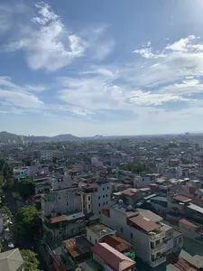 Bức ảnh chụp toàn cảnh một khu vực đô thị đông đúc, có thể là thành phố Thanh Hóa, nhìn từ trên cao. Khung cảnh bao gồm vô số các tòa nhà và nhà ở san sát nhau, với những mái nhà đa dạng về màu sắc và chất liệu như ngói đỏ và tôn. Trên nóc nhiều ngôi nhà có lắp đặt bồn chứa nước. Ở phía xa là những dãy đồi núi. Bầu trời trong xanh với những đám mây trắng. Ở phía dưới bên trái, có thể nhận ra một con đường với cây xanh, xe cộ đang đỗ và một vài bóng người. Không có chữ viết hay văn bản nào có thể đọc được trong ảnh.。（日本語訳）。上から見た、混雑した都市部、おそらくタインホア市のパノラマ写真です。風景には、無数のビルや住宅が密集しており、屋根は赤い瓦やトタンなど、色や素材も様々です。多くの家の屋上には貯水タンクが設置されています。遠くには丘や山々の連なりがあります。空は青く澄んでおり、白い雲が浮かんでいます。左下には、緑樹のある道、駐車している車両、そしていくつかの人影が見て取れます。写真の中には読み取れる文字やテキストはありません。