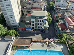 Bức ảnh chụp từ trên cao một góc phố đô thị ở Việt Nam, cho thấy một khu dân cư và thương mại. Các biển hiệu có thể đọc được bao gồm: 'NHÀ NGHỈ THU HÀ', cửa hàng 'AUMART' với dòng chữ 'CHUYÊN SỬA NỘI - SỮA NGOẠI - UY TÍN - CHẤT LƯỢNG', và các biển quảng cáo 'ĐÔNG TRÙNG HẠ THẢO Yến Sào Nguyên Tổ' và 'SẢN XUẤT XÚC XÍCH'. Khung cảnh mô tả hoạt động sinh hoạt thường ngày với người đi bộ, một số đội nón lá, và người dân di chuyển bằng xe máy trên đường. Một chiếc ô tô đang đỗ trước nhà nghỉ. Các vật thể nổi bật bao gồm nhiều tòa nhà cao tầng kiểu nhà ống san sát, hệ thống dây điện chằng chịt, và ở phía trước là một bể bơi ngoài trời với nước trong xanh, có ghế dài và ô che nắng.。（日本語訳）。ベトナムのある都市の街角を上空から撮影した写真で、住宅街と商業地区が写っている。読み取れる看板には、「NHÀ NGHỈ THU HÀ」（トゥーハー宿）、「CHUYÊN SỬA NỘI - SỮA NGOẠI - UY TÍN - CHẤT LƯỢNG」（国産粉ミルク・外国産粉ミルク専門 - 信頼 - 品質）と書かれた「AUMART」店、そして「ĐÔNG TRÙNG HẠ THẢO Yến Sào Nguyên Tổ」（冬虫夏草 ツバメの巣）と「SẢN XUẤT XÚC XÍCH」（ソーセージ製造）の広告看板が含まれる。この光景は、歩行者、ノンラー（笠）をかぶった人々、そして道路をバイクで移動する人々といった日常の生活活動を描写している。宿の前には一台の車が駐車している。