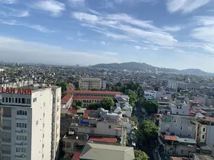 Đây là một bức ảnh chụp toàn cảnh một khu vực đô thị đông đúc, có khả năng là thành phố Thanh Hóa, được chụp từ trên cao vào một ngày nắng đẹp. Bầu trời trong xanh với những đám mây trắng. Ở phía tiền cảnh bên trái là một tòa nhà cao tầng màu trắng, trên nóc có biển hiệu chữ nổi màu đỏ ghi "LAN ANH" và các bồn chứa nước bằng inox. Nhìn bao quát là vô số các tòa nhà và nhà ở dân dụng với nhiều kiểu kiến trúc và mái nhà khác nhau (mái ngói đỏ, mái tôn, mái bằng) nằm san sát nhau. Xen kẽ giữa các công trình là cây xanh và những con đường nhỏ có xe cộ qua lại. Ở phía xa, có một ngọn đồi được bao phủ bởi cây xanh và trên đỉnh đồi có một cột ăng-ten hoặc tháp viễn thông.。（日本語訳）。これは、晴れた日に上空から撮影された、混雑した都市部、おそらくタインホア市のパノラマ写真です。空は青く澄んでおり、白い雲が浮かんでいます。左手前の前景には白い高層ビルがあり、その屋上には「LAN ANH」と書かれた赤い立体文字の看板とステンレス製の貯水タンクがあります。全体を見渡すと、様々な建築様式と異なる屋根（赤い瓦屋根、トタン屋根、陸屋根）を持つ無数のビルや住宅が密集しています。建物の間には緑の木々や、車が往来する小さな道が点在しています。遠くには、緑の木々で覆われた丘があり、その頂上にはアンテナまたは通信塔があります。