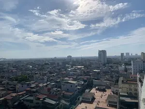 Đây là một bức ảnh chụp toàn cảnh một thành phố nhìn từ trên cao vào một ngày nắng đẹp. Bầu trời trong xanh với những đám mây trắng. Phía dưới là một khu đô thị đông đúc với vô số các tòa nhà và nhà ở san sát nhau. Các ngôi nhà ở gần chủ yếu là nhà thấp tầng, nhà ống với các mái nhà đa dạng và nhiều bồn chứa nước trên nóc. Ở phía xa, một vài tòa nhà chung cư và văn phòng cao tầng hiện đại nổi bật trên nền trời. Có thể thấy chữ viết trên một trong những tòa nhà cao tầng nhưng không thể đọc được nội dung. Khung cảnh tổng thể là một đô thị đang phát triển, không có hoạt động cụ thể nào của con người được ghi lại.。（日本語訳）。これは、晴れた美しい日に上空から見た都市の全景を撮影した写真です。空は青く澄み渡り、白い雲が浮かんでいます。眼下には、無数のビルや住宅が密集する、にぎやかな市街地が広がっています。手前にある家々は、主に低層住宅やチューブハウスで、屋根の形は様々で、屋上には多くの貯水タンクが設置されています。遠くには、近代的な高層マンションやオフィスビルがいくつか、空を背景に際立っています。高層ビルの一つに文字が見えますが、その内容は読み取れません。全体的な光景は発展途上の都市であり、特定の人間活動は写っていません。