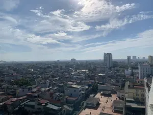 Đây là một bức ảnh chụp toàn cảnh một thành phố từ trên cao vào một ngày nắng đẹp. Bầu trời trong xanh với những đám mây trắng rải rác và ánh nắng mặt trời chói chang. Khung cảnh bên dưới là một khu đô thị dày đặc với vô số các tòa nhà và nhà ở san sát nhau. Ở phía trước, có thể thấy rõ các mái nhà của những ngôi nhà thấp tầng, bao gồm mái ngói, mái tôn và mái bằng bê tông. Trên một số mái nhà có lắp đặt bồn chứa nước và cục nóng điều hòa. Xen kẽ giữa các công trình xây dựng là một vài cây xanh. Ở phía xa hơn là các tòa nhà cao tầng hiện đại. Có chữ viết trên một tòa nhà cao tầng màu trắng ở phía bên phải nhưng không thể đọc được. Toàn cảnh cho thấy một thành phố đang phát triển, có thể là thành phố Thanh Hóa.。（日本語訳）。これは、晴れた美しい日に上空から都市の全景を撮影した写真です。空は青く澄み渡り、白い雲が点在し、太陽の光が眩しく輝いています。眼下には、無数のビルや住宅が密集する都市部が広がっています。手前には、瓦屋根、トタン屋根、コンクリートの陸屋根など、低層住宅の屋根がはっきりと見えます。いくつかの屋根の上には、貯水タンクやエアコンの室外機が設置されています。建物の間には、緑の木々がいくつか点在しています。さらに遠くには、近代的な高層ビルがあります。右側にある白い高層ビルには文字が書かれていますが、読むことはできません。全景は、発展途上にある都市、おそらくタインホア市であることを示しています。