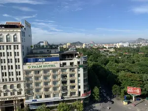 Đây là hình ảnh chụp toàn cảnh một khu vực đô thị từ trên cao, có thể là trung tâm thành phố Thanh Hóa.。1. Các dòng chữ có thể đọc được:。- Trên các tòa nhà: "ACB" và "NGÂN HÀNG Á CHÂU", "PALM HOTEL", "VIET HOTEL", "THANH HOA GROUP", "DAIVIET PALACE", và một biển hiệu màu đỏ có chữ "NHA SACH...".。- Trên một tấm biển quảng cáo lớn màu đỏ ở phía dưới bên phải: "GENERALI", "TẬP ĐOÀN BẢO HIỂM SỐ 1 TẠI Ý", "#SốngNhưÝ".。2. Mô tả cảnh quan và hoạt động:。- Loại địa điểm: Quang cảnh thành phố với nhiều tòa nhà cao tầng, khách sạn, văn phòng công ty và đường phố. Xung quanh có nhiều cây xanh và phía xa là những dãy núi.。- Hoạt động cụ thể: Trên đường phố bên dưới, có một vài chiếc ô tô và xe máy đang lưu thông. Đây là hoạt động giao thông thường ngày của một thành phố.。- Các vật thể trong ảnh: Các tòa nhà cao tầng với kiến trúc hiện đại, biển hiệu của các khách sạn và công ty, một biển quảng cáo lớn, cây xanh, ô tô, xe máy, và một bốt bảo vệ nhỏ màu vàng. Bầu trời trong xanh, có mây trắng.。（日本語訳）。これは上空から撮影した都市部の全景写真で、おそらくタインホア市の中心部です。1. 読み取れる文字：。- 建物の上：「ACB」と「NGÂN HÀNG Á CHÂU（アジア商業銀行）」、「PALM HOTEL」、「VIET HOTEL」、「THANH HOA GROUP」、「DAIVIET PALACE」、そして「NHA SACH...」という文字のある赤い看板。- 右下にある大きな赤い広告看板の上：「GENERALI」、「TẬP ĐOÀN BẢO HIỂM SỐ 1 TẠI Ý（イタリアNo.1の保険グループ）」、「#SốngNhưÝ」。2. 風景と活動の説明：。- 場所の種類：多くの高層ビル、ホテル、会社のオフィス、そして通りがある街の風景。周囲には多くの緑樹があり、遠くには山脈が見えます。- 具体的な活動：下の通りでは、数台の自動車とバイクが通行しています。これは都市の日常的な交通活動です。- 写真の中の物体：近代的な建築の高層ビル、ホテルや会社の看板、大きな広告看板、緑樹、自動車、バイク、そして小さな黄色の警備員詰所。空は青く澄んでおり、白い雲があります。