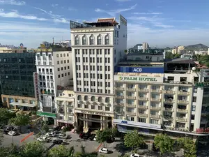 Đây là một bức ảnh chụp quang cảnh thành phố từ trên cao, có thể là thành phố Thanh Hóa, vào một ngày nắng đẹp, trời trong xanh. Trung tâm của bức ảnh là các tòa nhà cao tầng san sát nhau. Các văn bản và biển hiệu có thể nhận dạng bao gồm: "DAI VIET HOTEL", "PALM HOTEL", "ACB" (Ngân hàng Á Châu), "OCB" (Ngân hàng Phương Đông), "SeABank", "VietinBank", và "HOA SEN SPA". Ngoài ra còn có một biển hiệu dọc màu đỏ có chữ "MASSAGE" và một phần biển hiệu "Welcome to Palm Hotel". Dưới đường phố, có nhiều ô tô đang đỗ và cây xanh được trồng dọc hai bên. Một băng rôn màu đỏ được treo ngang qua đường nhưng không thể đọc rõ nội dung. Ở phía xa, có thể nhìn thấy những ngọn núi và các tòa nhà khác của thành phố.。（日本語訳）。これは、晴れた美しい日、青く澄んだ空の下、上空から撮影された街の風景写真で、おそらくタインホア市だと思われます。写真の中心には高層ビルが密集しています。認識できる文字や看板には、「DAI VIET HOTEL」、「PALM HOTEL」、「ACB」（アジア商業銀行）、「OCB」（オリエント商業銀行）、「SeABank」、「VietinBank」、そして「HOA SEN SPA」が含まれます。その他にも、「MASSAGE」と書かれた縦長の赤い看板や、「Welcome to Palm Hotel」という看板の一部があります。下の通りには、多くの車が駐車しており、両脇には緑の木々が植えられています。赤い横断幕が通りを横切って掲げられていますが、内容ははっきりと読み取れません。遠くには、山々や街の他の建物が見えます。