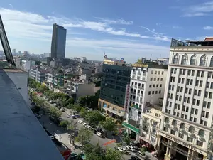 Bức ảnh được chụp từ trên cao, nhìn xuống một con phố chính sầm uất của một thành phố tại Việt Nam. Khung cảnh cho thấy một khu đô thị phát triển với nhiều tòa nhà cao tầng, bao gồm một tòa nhà chọc trời bằng kính hiện đại ở phía xa. Bầu trời trong xanh với vài gợn mây.。Hoạt động chính là giao thông đô thị, với nhiều xe ô tô và xe máy đang di chuyển và đậu dọc hai bên đường. Dọc con phố có nhiều cây xanh được trồng.。Nhiều biển hiệu của các doanh nghiệp có thể được nhìn thấy rõ, cho thấy đây là một trung tâm thương mại và dịch vụ. Các văn bản có thể đọc được bao gồm:。- Tên khách sạn: 'DAI VIET HOTEL'.。- Tên các ngân hàng: 'VietinBank', 'AGRIBANK', 'OCB' (với dòng chữ 'Ngân Hàng Phương Đông'), 'SeABank', 'eABank'.。- Tên dịch vụ: 'HOA SEN SPA', 'MASSAGE'.。- Một băng rôn màu đỏ treo ngang đường có các chữ như 'SỰ KIỆN VÀ ẨM THỰC', 'MAI', 'HỘI NGHỊ', 'GHIỆP', có thể là quảng cáo cho một sự kiện hoặc trung tâm hội nghị.。（日本語訳）。写真は上空から撮影されたもので、ベトナムのある都市の賑やかなメインストリートを見下ろしています。風景は、遠くにある近代的なガラス張りの超高層ビルを含む、多くの高層ビルが建ち並ぶ発展した都市部を示しています。空は青く澄んでおり、いくつかの雲が浮かんでいます。主な活動は都市交通で、多くの自動車やバイクが移動したり、道路の両側に駐車したりしています。通りに沿って多くの緑の木が植えられています。多くの企業の看板がはっきりと見え、ここが商業とサービスの中心地であることを示しています。読み取れるテキストには以下が含まれます：。- ホテル名：「DAI VIET HOTEL」。- 銀行名：「VietinBank」、「AGRIBANK」、「OCB」（「Ngân Hàng Phương Đông」の文字入り）、「SeABank」、「eABank」。- サービス名：「HOA SEN SPA」、「MASSAGE」。- 道路を横切って掛けられた赤い横断幕には、「SỰ KIỆN VÀ ẨM THỰC」、「MAI」、「HỘI NGHỊ」、「GHIỆP」などの文字があり、イベントや会議センターの広告である可能性があります。