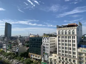 Bức ảnh chụp toàn cảnh một khu vực đô thị từ trên cao vào một ngày nắng đẹp, với bầu trời trong xanh và những gợn mây trắng. Khung cảnh cho thấy sự pha trộn giữa các tòa nhà hiện đại và các công trình kiến trúc cũ hơn. Nổi bật ở phía bên trái là một tòa nhà chọc trời hiện đại bằng kính. Ở trung tâm và phía bên phải là nhiều tòa nhà cao tầng khác. Có thể nhận dạng được một số biển hiệu của các doanh nghiệp như: 'VietinBank' trên nóc một tòa nhà, 'HOA SEN SPA' với chữ màu vàng, ngân hàng 'OCB', 'SeABank', và một biểu ngữ quảng cáo có chữ 'LOTTERIA'. Một tòa nhà lớn màu trắng ở phía trước bên phải có kiến trúc cổ điển với các cửa sổ hình vòm. Phía dưới bên trái là một con đường có nhiều cây xanh và ô tô đang đỗ, cho thấy một cảnh quan đô thị sầm uất.。（日本語訳）。晴れた日に上空から都市部を撮影した全景写真で、青い空と白い雲の筋がある。この風景は、現代的な建物とより古い建築物が混在している様子を示している。左側で目立っているのは、ガラス張りの現代的な超高層ビルである。中央と右側には、他の多くの高層ビルがある。ある建物の屋上にある「VietinBank」、黄色い文字の「HOA SEN SPA」、「OCB」銀行、「SeABank」、そして「LOTTERIA」という文字が入った広告バナーといった、いくつかの企業の看板が識別できる。右手前にある大きな白い建物は、アーチ型の窓を持つ古典的な建築様式である。左下には多くの緑の木々と駐車している車がある道があり、賑やかな都市景観を示している。