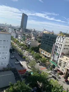 Bức ảnh chụp toàn cảnh một góc phố sầm uất từ trên cao, có thể là tại thành phố Thanh Hóa. Khung cảnh cho thấy một con đường lớn với nhiều ô tô đang di chuyển và đậu ven đường, hai bên là các tòa nhà cao tầng hiện đại và cây xanh. Bầu trời trong xanh, có nắng. Đây là một khu vực trung tâm thương mại với sự hiện diện của nhiều trụ sở ngân hàng và cơ sở kinh doanh.。Các vật thể và hoạt động chính bao gồm:。- **Địa điểm:** Một con đường đô thị lớn với các tòa nhà cao tầng, bao gồm một tòa nhà chọc trời bằng kính nổi bật.。- **Hoạt động:** Giao thông đường bộ với ô tô và xe máy. Các hoạt động thương mại diễn ra trong các tòa nhà.。- **Vật thể:** Ô tô, xe máy, cây xanh, cột điện, biển báo và các tòa nhà văn phòng, cửa hàng.。Thông tin chữ viết có thể nhận dạng được trên các biển hiệu:。- **Ngân hàng:** "VietinBank", "SeABank", "OCB" (Ngân Hàng Phương Đông), "Agribank".。- **Cơ sở kinh doanh khác:** "HOA SEN SPA", "MASSAGE", "LOTTERIA".。- **Băng rôn quảng cáo:** Một tấm biển lớn màu đỏ quảng cáo cho "TRUNG TÂM TỔ CHỨC SỰ KIỆN VÀ ẨM THỰC SAO MAI", chuyên về "SỰ KIỆN, TIỆC CƯỚI, HỘI NGHỊ" với khẩu hiệu "TRỌNG - CHUYÊN NGHIỆP". Một số băng rôn khác treo ngang đường nhưng không đọc rõ chữ.。（日本語訳）。上空から撮影された、賑やかな街角の全景写真で、おそらくタインホア市で撮影されたものでしょう。空は青く晴れ渡り、日差しがあります。ここは多くの銀行本店や商業施設が集まる商業中心地です。主な物体と活動は以下の通りです：。- **場所：** 高層ビルが立ち並ぶ都市の大きな道路で、ひときわ目立つガラス張りの超高層ビルが含まれます。- **活動：** 自動車やバイクによる道路交通。ビル内で行われる商業活動。- **物体：** 自動車、バイク、緑の木々、電柱、看板、そしてオフィスビルや店舗。看板で識別できる文字情報は以下の通りです：。- **銀行：** 「VietinBank」、「SeABank」、「OCB」（東方銀行）、「Agribank」。- **その他の商業施設：** 「HOA SEN SPA」、「MASSAGE」、「LOTTERIA」。- **広告バナー：** 「TRUNG TÂM TỔ CHỨC SỰ KIỆN VÀ ẨM THỰC SAO MAI」（サオマイ イベント・飲食センター）の大きな赤い看板があり、「イベント、結婚披露宴、会議」を専門とし、「尊重 - プロフェッショナル」というスローガンが掲げられています。その他にも道路を横切っていくつかのバナーが掲げられていますが、文字ははっきりと読み取れません。