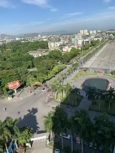 Bức ảnh chụp một khung cảnh đô thị từ trên cao, có thể là thành phố Thanh Hóa, vào một ngày nắng đẹp.。**Địa điểm và cảnh vật:**。*   **Quảng trường và đường phố:** Trung tâm của bức ảnh là một quảng trường công cộng rất lớn, được lát đá, bên cạnh là một đại lộ. Dọc theo đường và quảng trường được trồng nhiều hàng cây cọ. Trên quảng trường có một đài phun nước hình tròn hiện không hoạt động và một số công trình trang trí như tác phẩm điêu khắc hình trái tim màu đỏ.。*   **Công trình xây dựng:** Cảnh quan thành phố bao gồm nhiều loại nhà ở, các tòa nhà thương mại và một vài tòa nhà chung cư cao tầng ở phía xa.。*   **Thiên nhiên:** Khu vực có rất nhiều cây xanh, bao gồm cây cọ, cây phượng vĩ đang nở hoa đỏ. Ở phía xa là dãy núi trập trùng dưới bầu trời xanh.。**Hoạt động:**。*   Giao thông trên đường diễn ra bình thường với một vài chiếc xe máy và ô tô đang di chuyển. Quảng trường khá vắng vẻ, không có hoạt động công cộng nào đang diễn ra.。**Chữ viết:**。*   Một biển quảng cáo lớn màu đỏ của công ty bảo hiểm GENERALI có thể được nhìn thấy, với chữ "BẢO HIỂM SỐ 1".。*   Có một cổng chào trang trí bắc qua một con đường. Chữ trên cổng chào khó đọc, nhưng dường như là một thông điệp chào mừng, có thể liên quan đến một phường địa phương (Phường Lam Sơn).。*   Một tấm biển nhỏ ghi chữ "MASSAGE" được nhìn thấy ở phía dưới.。（日本語訳）。写真は、ある晴れた日に上空から撮影された都市の風景で、おそらくタインホア市だと思われます。**場所と景色：**。*   **広場と通り：**写真の中心には、石畳で舗装された非常に大きな公共広場があり、その隣には大通りがあります。通りと広場に沿って、多くのヤシの木が植えられています。広場には、現在は稼働していない円形の噴水と、赤いハート形の彫刻作品のような装飾的な建造物がいくつかあります。*   **建造物：**都市の景観には、多くの種類の住宅、商業ビル、そして遠くにはいくつかの高層マンションが含まれています。*   **自然：**この地域には、ヤシの木や赤い花を咲かせているホウオウボクなど、多くの緑があります。遠くには、青空の下に連なる山々が見えます。**活動：**。*   道路の交通は通常通りで、数台のバイクと車が移動しています。広場はかなり閑散としており、公共の活動は行われていません。**文字：**。*   GENERALI保険会社の大きな赤い広告看板が見え、「BẢO HIỂM SỐ 1」（ナンバーワン保険）という文字が書かれています。*   ある道をまたぐように装飾的な歓迎ゲートがあります。ゲートの文字は読みにくいですが、歓迎のメッセージのようで、地元の区（ラムソン区）に関連している可能性があります。*   下の方に「MASSAGE」と書かれた小さな看板が見えます。