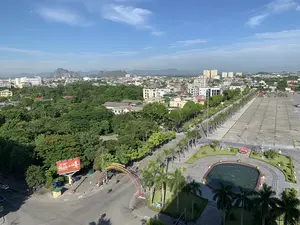 Bức ảnh chụp toàn cảnh một khu vực đô thị từ trên cao vào một ngày nắng đẹp, trời trong xanh. Khung cảnh bao gồm một quảng trường công cộng rộng lớn, các con đường, nhiều cây xanh và các tòa nhà. Về chữ viết, có một cổng chào trang trí bắc qua đường với dòng chữ màu vàng: 'PHƯỜNG ĐÔNG HƯƠNG CHÀO MỪNG QUÝ KHÁCH' và bên dưới là 'KHU PHỐ VĂN MINH - QUẢNG TRƯỜNG LAM SƠN'. Ngoài ra còn có một biển quảng cáo lớn màu đỏ của 'GENERALI' với nội dung 'SỐ 1 VỀ BẢO HIỂM SỐ TRỊ GIÁ' và hashtag '#SốngNhưÝ'. Về cảnh vật và hoạt động: Địa điểm là một thành phố, cụ thể là khu vực xung quanh một quảng trường lớn (Quảng trường Lam Sơn) và một trục đường chính. Phía xa có thể thấy những dãy núi đá vôi. Hoạt động chính là giao thông trên đường khá thưa thớt, chủ yếu là xe máy và một vài chiếc ô tô đang di chuyển. Quảng trường gần như trống không, không có sự kiện nào đang diễn ra. Các vật thể nổi bật bao gồm quảng trường lát gạch rộng lớn, một đài phun nước hình bầu dục (không hoạt động), các hàng cây cọ được trồng thẳng tắp dọc theo đường và quảng trường. Trong khu vực cây xanh của quảng trường có một công trình trang trí hình trái tim màu đỏ. Các tòa nhà đa dạng từ nhà dân thấp tầng đến các tòa nhà cao tầng.。（日本語訳）。晴れた美しい日、青空の下、上空から都市部を撮影した全景写真です。風景には、広大な公共広場、道路、多くの緑樹、そして建物が含まれています。文字については、道路をまたぐ装飾的な歓迎ゲートがあり、黄色い文字で「PHƯỜNG ĐÔNG HƯƠNG CHÀO MỪNG QUÝ KHÁCH」（ドンフオン区は皆様を歓迎します）と書かれ、その下には「KHU PHỐ VĂN MINH - QUẢNG TRƯỜNG LAM SƠN」（文明的な街区 - ラムソン広場）とあります。さらに、「GENERALI」の大きな赤い広告看板があり、「SỐ 1 VỀ BẢO HIỂM SỐ TRỊ GIÁ」（価値あるデジタル保険でNo.1）という内容とハッシュタグ「#SốngNhưÝ」（思い通りの人生を）が書かれています。風景と活動について：場所は都市で、具体的には大きな広場（ラムソン広場）と主要な幹線道路の周辺地域です。遠くには石灰岩の山々が見えます。主な活動は道路上の交通で、かなりまばらで、主にバイクと数台の自動車が移動しています。広場はほとんど誰もおらず、何のイベントも開催されていません。目立つ物体には、広大なタイル張りの広場、楕円形の噴水（稼働していない）、道路と広場に沿ってまっすぐに植えられたヤシの木の列が含まれます。広場の緑地エリアには、赤いハート型の装飾物があります。建物は低層の民家から高層ビルまで様々です。