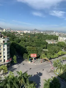 Đây là quang cảnh thành phố Thanh Hóa được chụp từ trên cao vào một ngày nắng đẹp, trời trong xanh. Phía trước là một ngã tư đường phố với một vài người đang điều khiển xe máy. Xung quanh có rất nhiều cây xanh, bao gồm cả cây cọ, xen kẽ giữa các tòa nhà. Về phía bên trái, có một tòa nhà cao tầng với biển hiệu "THANH HOA GROUP". Ở trung tâm, có một biển quảng cáo lớn màu đỏ của hãng bảo hiểm "GENERALI" với dòng chữ "BẢO HIỂM SỐ 1". Một cổng chào trang trí bắc qua đường có dòng chữ: "NHIỆT LIỆT CHÀO MỪNG CÁC VẬN ĐỘNG VIÊN VỀ THAM DỰ GIẢI BÓNG BÀN CÁC CLB THÀNH PHỐ THANH HÓA - CÚP BIA THANH HÓA...". Phía xa là khung cảnh thành phố với nhiều nhà cửa và những dãy núi đá vôi đặc trưng của vùng. Có thể nhận ra một tòa nhà của "DAI HOC HONG DUC" (Đại học Hồng Đức) ở phía xa.。（日本語訳）。これは、晴れた美しい日、青空の下で上空から撮影されたタインホア市の光景です。手前には交差点があり、数人がバイクを運転しています。周囲にはヤシの木を含む多くの緑の木々が、建物の間に点在しています。左側には、「THANH HOA GROUP」という看板のある高層ビルがあります。中央には、「BẢO HIỂM SỐ 1」（ナンバーワン保険）という文字が書かれた保険会社「GENERALI」の大きな赤い広告看板があります。道路をまたぐ装飾された歓迎ゲートには、「NHIỆT LIỆT CHÀO MỪNG CÁC VẬN ĐỘNG VIÊN VỀ THAM DỰ GIẢI BÓNG BÀN CÁC CLB THÀNH PHỐ THANH HÓA - CÚP BIA THANH HÓA...」（タインホア市クラブ卓球大会 - タインホアビールカップ...に参加する選手たちを熱烈に歓迎します）という文字があります。遠方には、多くの家々やこの地域特有の石灰岩の山々が連なる街の風景が広がっています。遠くに「DAI HOC HONG DUC」（ホンĐức大学）の建物が確認できます。