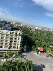 Đây là một bức ảnh chụp toàn cảnh một góc thành phố từ trên cao, có khả năng là thành phố Thanh Hóa, vào một ngày nắng đẹp.。**Chữ viết được ghi lại:**。*   Trên tòa nhà cao tầng bên trái: 'ACB', '...M HOTEL', 'THANH... GROUP', 'NGÂN HÀNG Á CHÂU', 'Welcome to Palm Hotel', và một biển hiệu màu đỏ có chữ 'NHA KHOA...' nhưng bị che một phần.。*   Trên biển quảng cáo lớn màu đỏ ở bên phải: logo 'GENERALI', 'BẢO HIỂM SỐ 1 TẠI Ý', và '#SốngNhưÝ'.。*   Trên cổng chào trang trí: Có các chữ màu vàng nhưng không thể đọc rõ toàn bộ nội dung.。**Mô tả cảnh vật và hoạt động:**。*   **Loại địa điểm:** Đây là một quang cảnh đô thị, nhìn xuống một ngã ba đường phố từ một tòa nhà cao. Xung quanh là các tòa nhà thương mại, khu dân cư, và rất nhiều cây xanh. Phía xa có những dãy núi đá vôi đặc trưng.。*   **Hoạt động:** Giao thông trên đường khá thưa thớt, có một vài chiếc xe máy đang chạy và một số ô tô đang đỗ. Cảnh vật thể hiện một buổi sáng hoặc chiều yên tĩnh trong thành phố.。*   **Vật thể trong ảnh:** Tòa nhà chính trong ảnh là một khách sạn lớn. Ngoài ra còn có các tòa nhà khác, đường nhựa, ô tô, xe máy, cây cọ, cây phượng, biển quảng cáo, và một cổng chào. Trên nóc các tòa nhà có thể thấy các thiết bị như chảo vệ tinh và dàn nóng điều hòa.。（日本語訳）。これは、晴れた美しい日に、上空から撮影された街の一角のパノラマ写真で、おそらくタインホア市だと思われます。**記録された文字：**。*   左側の高層ビルには、「ACB」、「...M HOTEL」、「THANH... GROUP」、「NGÂN HÀNG Á CHÂU（アジア商業銀行）」、「Welcome to Palm Hotel」、そして「NHA KHOA...」という文字がある赤い看板がありますが、一部が隠れています。*   右側の大きな赤い広告看板には、「GENERALI」のロゴ、「BẢO HIỂM SỐ 1 TẠI Ý（イタリアNo.1の保険）」、そして「#SốngNhưÝ（思い通りに生きよう）」と書かれています。**風景と活動の説明：**。*   **場所の種類：** これは都市の風景で、高い建物から三叉路を見下ろしています。周囲には商業ビル、住宅地、そして多くの緑の木々があります。遠くには特徴的な石灰岩の山脈が見えます。*   **活動：** 道路の交通量はかなり少なく、数台のバイクが走行しており、いくつかの車が駐車しています。この風景は、街の静かな朝または午後を表しています。*   **写真の中の物体：** 写真の主要な建物は大きなホテルです。その他にも、他の建物、アスファルトの道路、自動車、バイク、ヤシの木、ホウオウボク、広告看板、そして歓迎ゲートがあります。建物の屋上には、衛星放送受信アンテナやエアコンの室外機などの設備が見えます。