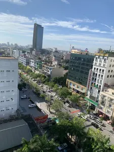 Bức ảnh chụp từ trên cao một góc phố của một thành phố, có thể là thành phố Thanh Hóa, vào một ngày nắng đẹp, trời xanh và có mây trắng. Khung cảnh cho thấy một con đường lớn với nhiều cây xanh hai bên. Dọc con đường là các tòa nhà cao tầng san sát, bao gồm cả một tòa nhà chọc trời hiện đại ở phía xa. Giao thông trên đường khá tấp nập với nhiều ô tô và xe máy đang di chuyển và đậu ven đường. Có thể nhận diện được nhiều biển hiệu của các doanh nghiệp và ngân hàng, cho thấy đây là một khu vực thương mại sầm uất.。Các dòng chữ có thể đọc được bao gồm:。- Biển hiệu ngân hàng: 'VietinBank', 'OCB Ngân Hàng Phương Đông', 'SeABank'.。- Biển hiệu dịch vụ: 'HOA SEN SPA', và một biển hiệu dọc màu đỏ có chữ 'MASSAGE'.。- Một tấm băng rôn quảng cáo lớn màu đỏ và vàng có nội dung: 'TRUNG TÂM TỔ CHỨC SỰ KIỆN VÀ ẨM THỰC SAO MAI', bên dưới là 'TỔ CHỨC SỰ KIỆN, TIỆC CƯỚI, HỘI NGHỊ' và 'TRANG TRỌNG - CHUYÊN NGHIỆP'.。Ngoài ra còn có các băng rôn khác treo ngang đường nhưng không đọc rõ chữ.。（日本語訳）。ある都市、おそらくタインホア市の街角を上空から撮影した写真。晴れた美しい日で、空は青く、白い雲が浮かんでいる。風景には、両側に多くの緑の木々が植えられた大きな通りが写っている。道路の交通はかなり混雑しており、多くの自動車やバイクが走行したり、道端に駐車したりしている。多くの企業や銀行の看板が認識でき、ここが賑やかな商業地域であることを示している。読み取れる文字は以下の通り：。- 銀行の看板：「VietinBank」、「OCB東方銀行」、「SeABank」。- サービスの看板：「HOA SEN SPA」、そして「MASSAGE」という文字が入った赤い縦長の看板。- 赤と黄色の大きな広告バナーには、「SAO MAIイベント・飲食センター」と書かれ、その下には「イベント、結婚披露宴、会議の開催」そして「格調高く - プロフェッショナルに」とある。