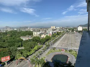 Bức ảnh chụp toàn cảnh một góc thành phố Thanh Hóa từ trên cao vào một ngày đẹp trời, bầu trời trong xanh và có ít mây. Vị trí trung tâm của bức ảnh là Quảng trường Lam Sơn, một không gian công cộng rộng lớn được lát đá. Trên quảng trường có một đài phun nước hình bầu dục (hiện không hoạt động) và một tác phẩm điêu khắc hình trái tim màu đỏ. Bên cạnh quảng trường là một con đường lớn rợp bóng cây xanh, bao gồm cả những hàng cây cọ. Giao thông trên đường khá thưa thớt với một vài chiếc ô tô và xe máy. Nhìn ra xa là khu đô thị với nhiều tòa nhà cao tầng, nhà dân xen lẫn với những mảng cây xanh rậm rạp. Ở phía chân trời là những dãy núi đá vôi. Có thể đọc được một số văn bản trong ảnh: một biển quảng cáo lớn màu đỏ của công ty bảo hiểm GENERALI với khẩu hiệu 'BẢO HIỂM SỐ 1 TẠI Ý' và '#SốngNhưÝ'; và một băng rôn trên cổng chào bắc qua đường ghi 'PHỐ ĐI BỘ PHAN CHU TRINH' và 'KHÔNG GIAN VĂN HÓA QUẢNG TRƯỜNG LAM SƠN'.。（日本語訳）。晴れた日に上空から撮影されたタインホア市の一角の全景写真で、空は青く澄み渡り、雲はほとんどありません。写真の中央には、石畳の広大な公共スペースであるラムソン広場があります。広場には、楕円形の噴水（現在は稼働していません）と、赤いハート形の彫刻作品があります。広場の隣には、ヤシの木を含む緑の木々が影を落とす大きな道路があります。道路の交通量はかなり少なく、数台の自動車とバイクが見られます。遠くには、多くの高層ビルや民家が鬱蒼とした緑地と混在する市街地が見えます。地平線には石灰岩の山々が連なっています。写真の中ではいくつかの文字を読むことができます。保険会社GENERALIの大きな赤い広告看板には、「BẢO HIỂM SỐ 1 TẠI Ý」（イタリアでNo.1の保険）と「#SốngNhưÝ」というスローガンが書かれています。また、道路をまたぐ歓迎ゲートの横断幕には、「PHỐ ĐI BỘ PHAN CHU TRINH」（ファン・チュー・チン歩行者天国）と「KHÔNG GIAN VĂN HÓA QUẢNG TRƯỜNG LAM SƠN」（ラムソン広場文化空間）と書かれています。