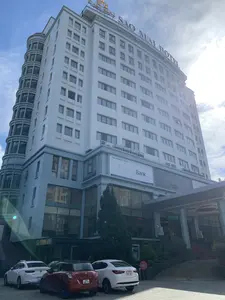 Bức ảnh chụp mặt tiền của một tòa nhà cao tầng hiện đại, đó là Khách sạn Sao Mai (Sao Mai Hotel) tại thành phố Thanh Hóa. Tòa nhà có màu sơn trắng và xanh nhạt, với nhiều cửa sổ và một phần kiến trúc hình trụ ở bên cạnh. Trên nóc tòa nhà có biển hiệu lớn ghi "SAO MAI HOTEL". Ở mặt tiền tòa nhà có một tấm biển quảng cáo lớn cho Ngân hàng Hợp tác xã Việt Nam (Co-opBank) với nội dung: "Ngân hàng Hợp tác xã Việt Nam - Co-opBank - Tầng 3 Tòa nhà Khách sạn Sao Mai - Số 20 Phan Chu Trinh - P. Điện Biên - TP. Thanh Hóa - www.co-opbank.vn". Lối vào chính của khách sạn có mái che, bên dưới có biển hiệu "SAO MAI HOTEL" và một bảng điện tử chữ chạy màu xanh lá nhưng không đọc rõ nội dung. Một cầu thang trải thảm đỏ dẫn lên sảnh chính. Phía trước tòa nhà là bãi đỗ xe, có một vài chiếc ô tô đang đỗ. Khung cảnh cho thấy đây là một ngày nắng, trời trong xanh.。（日本語訳）。近代的な高層ビルの正面を写した写真で、タインホア市にあるサオマイホテル（Sao Mai Hotel）です。建物は白と薄い青色に塗られており、多くの窓と、側面には円筒形の建築部分があります。建物の屋上には「SAO MAI HOTEL」と書かれた大きな看板があります。建物の正面には、ベトナム協同組合銀行（Co-opBank）の大きな広告看板があり、その内容は「ベトナム協同組合銀行 - Co-opBank - サオマイホテルビル3階 - タインホア市ディエンビエン区ファン・チュー・チン通り20番地 - www.co-opbank.vn」です。ホテルの正面玄関には屋根があり、その下には「SAO MAI HOTEL」という看板と、緑色の文字が流れる電光掲示板がありますが、内容ははっきりと読めません。赤いカーペットが敷かれた階段がメインロビーに通じています。建物の前は駐車場で、数台の車が駐車しています。風景は、これが晴れた日で、空が青く澄んでいることを示しています。