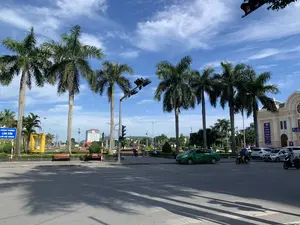 Bối cảnh là một góc phố tại một ngã tư ở thành phố Thanh Hóa vào một ngày nắng đẹp, trời trong xanh với mây trắng. Bên trái là Quảng trường Lam Sơn, được nhận diện qua tấm biển chỉ dẫn màu xanh ghi 'QUẢNG TRƯỜNG LAM SƠN' và 'LAM SON SQUARE'. Trong quảng trường có nhiều cây cọ cao, các công trình điêu khắc trang trí, ghế đá và một vài người đang ngồi. Trên đường phố, các phương tiện giao thông như một chiếc taxi màu xanh lá, ô tô con và xe máy đang di chuyển qua lại. Hệ thống đèn tín hiệu giao thông đang hoạt động tại ngã tư, và bóng của những hàng cây đổ dài trên mặt đường. Bên phải là một tòa nhà lớn với kiến trúc đặc trưng, là Nhà hát Lam Sơn. Trên tòa nhà có treo các biểu ngữ, trong đó có dòng chữ 'LIÊN HOAN PHÁT THANH TOÀN QUỐC' và 'CHÀO MỪNG'. Phía xa trong hậu cảnh có thể thấy các tòa nhà cao tầng. Hoạt động chính là giao thông đô thị và sinh hoạt công cộng tại quảng trường.。（日本語訳）。背景は、タインホア市のある交差点の街角で、空は青く澄み、白い雲が浮かぶ晴れた美しい日です。左側はラムソン広場で、「QUẢNG TRƯỜNG LAM SƠN」と「LAM SON SQUARE」と書かれた青い案内標識で識別できます。広場の中には、高いヤシの木がたくさんあり、装飾的な彫刻作品、石のベンチがあり、数人が座っています。路上では、緑色のタクシー、乗用車、バイクなどの車両が行き来しています。交差点では交通信号機が作動しており、並木道の影が路面に長く伸びています。右側は特徴的な建築様式の大きな建物で、ラムソン劇場です。建物には横断幕が掲げられており、その中には「LIÊN HOAN PHÁT THANH TOÀN QUỐC」と「CHÀO MỪNG」という文字があります。遠くの背景には高層ビルが見えます。主な活動は、都市交通と広場での公共の営みです。