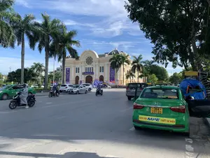 Bức ảnh chụp khung cảnh đường phố vào ban ngày tại thành phố Thanh Hóa, phía trước Nhà hát Lam Sơn. Tòa nhà nhà hát có kiến trúc cổ điển màu kem, trên mặt tiền có dòng chữ "NHÀ HÁT LAM SƠN". Phía trước tòa nhà được trang trí bằng các biểu ngữ màu xanh dương cho một sự kiện, với nội dung: "LIÊN HOAN PHÁT THANH TOÀN QUỐC LẦN THỨ XVI - 2024" và "CHÀO MỪNG ĐẠI BIỂU THAM DỰ LIÊN HOAN PHÁT THANH TOÀN QUỐC LẦN THỨ XVI - 2024". Trên đường có nhiều ô tô và xe máy đang di chuyển và đậu đỗ. Bên phải hình ảnh là một chiếc taxi màu xanh lá cây đang đỗ, trên thân xe có các dòng chữ "TỔNG ĐÀI TAXI TOÀN QUỐC 1055" và quảng cáo "ĐÈN LED MPE THIẾT BỊ ĐIỆN". Dọc theo con đường là những hàng cây cọ cao và cây xanh khác.。（日本語訳）。タインホア市、ラムソン劇場の前で日中に撮影された街の風景の写真。劇場の建物はクリーム色の古典的な建築様式で、正面には「NHÀ HÁT LAM SƠN」の文字がある。建物の前は、イベントのために青い横断幕で飾られており、その内容は「第16回全国放送フェスティバル - 2024」と「第16回全国放送フェスティバル - 2024に参加する代表者を歓迎します」である。路上には多くの自動車やバイクが走行したり駐車したりしている。写真の右側には緑色のタクシーが1台駐車しており、車体には「全国タクシーコールセンター 1055」の文字と「MPE LED照明 電気設備」の広告がある。道沿いには高いヤシの木やその他の緑の木々が並んでいる。