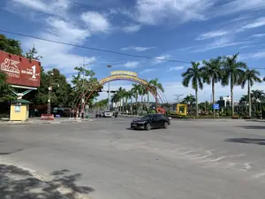 Bức ảnh chụp một ngã tư hoặc vòng xuyến lớn tại một thành phố vào một ngày nắng đẹp, trời trong xanh và có mây. Các phương tiện giao thông như ô tô và xe máy đang di chuyển trên đường.。Các thông tin bằng chữ có thể nhận dạng được:。- Trên cổng chào lớn màu đỏ và vàng bắc qua đường có dòng chữ: "PHỐ ĐI BỘ PHAN CHU TRINH" và "KHÔNG GIAN VĂN HÓA QUẢNG TRƯỜNG LAM SƠN".。- Ở bên trái, có một tấm biển quảng cáo lớn màu đỏ của công ty bảo hiểm Generali với nội dung: "GENERALI", "TẬP ĐOÀN BẢO HIỂM SỐ 1 TẠI Ý", và "#SốngNhưÝ".。- Ở phía xa hơn, có một biển báo giao thông màu xanh ghi "QUẢNG TRƯỜNG LAM SƠN" và "LAM SON SQUARE".。Cảnh vật và hoạt động:。- Địa điểm: Đây là một giao lộ đường phố, dẫn vào khu vực Phố đi bộ Phan Chu Trinh và Quảng trường Lam Sơn tại thành phố Thanh Hóa.。- Hoạt động: Các phương tiện đang tham gia giao thông. Một chiếc ô tô màu đen đang đi vào vòng xuyến, cùng với một vài xe máy và một chiếc ô tô màu trắng ở phía sau.。- Vật thể: Có một cổng chào lớn, một biển quảng cáo, một bốt gác nhỏ màu vàng, nhiều cây cọ được trồng dọc hai bên đường, cột đèn đường và đèn tín hiệu giao thông. Phía sau là không gian công cộng của quảng trường.。（日本語訳）。ある都市の大きな交差点またはロータリーを、晴れた日に撮影した写真。空は青く澄み、雲が浮かんでいる。自動車やバイクなどの交通車両が道路を走行している。認識可能な文字情報：。- 道路をまたぐ赤と黄色の大きな歓迎ゲートには、「PHỐ ĐI BỘ PHAN CHU TRINH」（ファン・チュー・チン歩行者天国）と「KHÔNG GIAN VĂN HÓA QUẢNG TRƯỜNG LAM SƠN」（ラムソン広場文化空間）という文字がある。- 左側には、ゼネラリ保険会社の大きな赤い広告看板があり、「GENERALI」、「TẬP ĐOÀN BẢO HIỂM SỐ 1 TẠI Ý」（イタリアNo.1保険グループ）、そして「#SốngNhưÝ」という内容が書かれている。- さらに奥には、「QUẢNG TRƯỜNG LAM SƠN」と「LAM SON SQUARE」と記された青い交通標識がある。風景と活動：。- 場所：これはタインホア市にあるファン・チュー・チン歩行者天国とラムソン広場のエリアに通じる道路の交差点である。- 活動：車両が交通に参加している。一台の黒い自動車がロータリーに進入しており、数台のバイクと後方の一台の白い自動車もいる。- 物体：大きな歓迎ゲート、広告看板、小さな黄色い警備詰所、道路の両脇に植えられた多くのヤシの木、街灯、そして信号機がある。後方は広場の公共スペースになっている。