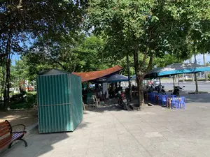 Đây là hình ảnh một quán nước vỉa hè, thường được gọi là quán cóc, nằm trên vỉa hè dưới bóng mát của những cây xanh lớn. Quán được che bằng những chiếc ô lớn nhiều màu sắc và tấm bạt. Có nhiều bộ bàn ghế nhựa nhỏ màu xanh và cam được bày ra cho khách ngồi. Một vài người đang ngồi uống nước và thư giãn. Gần đó có một số xe máy đang đỗ. Bên trái có một ki-ốt di động làm bằng tôn màu xanh lá cây đang đóng cửa. Phía sau là một con đường nhựa rộng rãi với xe cộ qua lại. Không có văn bản hay biển hiệu nào có thể đọc được trong ảnh.。（日本語訳）。これは、一般に「クアンコック」と呼ばれる路上カフェの写真で、大きな緑の木々の木陰の歩道にあります。店は、色とりどりの大きな傘と防水シートで覆われています。客が座るために、青色とオレンジ色の小さなプラスチック製のテーブルと椅子のセットが多数並べられています。数人が座って飲み物を飲み、くつろいでいます。近くには数台のバイクが駐車されています。左側には、緑色のトタンでできた移動式キオスクがあり、閉まっています。後方には、車両が行き交う広々とした舗装道路があります。写真には、読み取れる文字や看板はありません。