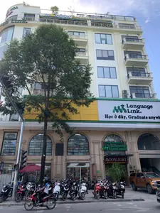 Bức ảnh chụp một tòa nhà cao tầng trên một con phố ở thành phố. Trên tòa nhà có nhiều biển hiệu với các dòng chữ: 'THANH HOA GROUP', 'moonlight Bistro Lounge & Restaurant' ở trên nóc; 'Language Link. Học ở đây, Graduate anywhere' ở các tầng giữa; và 'TRUNG TÂM ANH NGỮ Language Link ACADEMIC', 'MOONLIGHT Bistro Lounge & Restaurant', 'THANG MÁY AZ' ở tầng trệt.。Đây là một khu vực thương mại, có một trung tâm Anh ngữ và một nhà hàng. Phía trước tòa nhà, trên vỉa hè, có rất nhiều xe máy đang đậu. Một cây xanh lớn che một phần mặt tiền của tòa nhà. Có một cột đèn tín hiệu giao thông ở bên trái. Một người đàn ông đang lái xe máy trên đường, cho thấy hoạt động giao thông thường ngày. Ngoài ra còn có một chiếc ô tô bán tải màu cam đang đậu gần đó.。（日本語訳）。写真は、街の通りにある高層ビルを写しています。ビルには多くの看板があり、屋上には「THANH HOA GROUP」、「moonlight Bistro Lounge & Restaurant」という文字があります。中間階には「Language Link. ここで学び、どこでも卒業する（Học ở đây, Graduate anywhere）」とあります。そして1階には「英語センター Language Link ACADEMIC」、「MOONLIGHT Bistro Lounge & Restaurant」、「AZエレベーター」とあります。ここは商業エリアで、英語センターとレストランがあります。ビルの前、歩道にはたくさんのバイクが停まっています。大きな緑の木がビルの正面の一部を覆っています。左側には信号機があります。一人の男性が道路でバイクを運転しており、日常の交通活動を示しています。さらに、近くにはオレンジ色のピックアップトラックが停まっています。