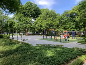 Bức ảnh chụp một không gian công cộng ngoài trời, có thể là một công viên hoặc một khu vườn hoa. Nổi bật trong ảnh là khu vui chơi dành cho trẻ em với nền cỏ nhân tạo màu xanh và các thiết bị vui chơi nhiều màu sắc như xích đu, cầu trượt, thú nhún và giàn leo trèo. Có một biển báo được dựng lên trong khu vực này, nhưng nội dung chữ viết không thể đọc rõ. Bên cạnh khu vui chơi là khu vực tập thể dục với nền sân lát gạch, được trang bị các dụng cụ thể thao ngoài trời. Xung quanh là rất nhiều cây xanh tươi tốt, bao gồm cả cây phượng có hoa đỏ. Phía sau, có thể thấy một bãi đỗ xe với một vài chiếc ô tô. Bầu trời trong xanh, quang đãng, cho thấy thời tiết đẹp. Khung cảnh chung rất yên tĩnh và không có người.。（日本語訳）。写真は屋外の公共スペースを写しており、公園か花園のようです。写真の中で目立っているのは、緑色の人工芝が敷かれた子供用の遊び場で、ブランコ、滑り台、スプリング遊具、ジャングルジムといった色とりどりの遊具があります。このエリアには看板が一つ設置されていますが、書かれている文字の内容ははっきりと読み取れません。遊び場の隣には、タイル張りの床に屋外運動器具が設置された運動エリアがあります。周りには、赤い花を咲かせたホウオウボクを含む、青々とした木々がたくさんあります。後方には、数台の車が停まっている駐車場が見えます。空は青く澄み渡っており、天気が良いことを示しています。全体的な光景はとても静かで、人はいません。