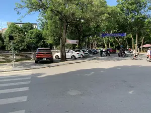 Bức ảnh chụp một con đường tại một thành phố ở Việt Nam, có thể là ở Thanh Hóa, vào một ngày nắng. Hai bên đường có nhiều cây xanh lớn tỏa bóng mát. Dưới lòng đường có vạch kẻ dành cho người đi bộ. Nhiều ô tô đang đỗ ở lề đường, trong đó có một chiếc SUV màu đỏ của hãng VinFast. Nhiều người đang điều khiển xe máy tham gia giao thông. Bên trái bức ảnh là một con kênh nhỏ có lan can bảo vệ. Có hai băng rôn được treo trên cây. Băng rôn màu xanh có nội dung: 'CHÀO MỪNG ĐẠI BIỂU THAM DỰ LIÊN HOAN PHÁT THANH TOÀN QUỐC LẦN THỨ XVI - 2024. Thanh Hóa, ngày 09 - 14/7/2024'. Một băng rôn khác có chữ 'GRAND OPENING 05.07.2024', dường như là quảng cáo cho một sự kiện khai trương.。（日本語訳）。この写真は、晴れた日にベトナムのある都市、おそらくタインホアの道路を撮影したものです。道の両側には、涼しい木陰を作る大きな緑の木がたくさんあります。路面には横断歩道の白線が引かれています。多くの車が路肩に駐車しており、その中にはVinFast（ビンファスト）社の赤いSUVが1台あります。多くの人々がバイクを運転して交通に参加しています。写真の左側には、手すりで保護された小さな水路があります。木には2つの横断幕が掲げられています。青い横断幕には、「第16回全国ラジオフェスティバル2024に参加する代表者を歓迎します。タインホア、2024年7月9日～14日」という内容が書かれています。もう一つの横断幕には「GRAND OPENING 05.07.2024」と書かれており、これは開店イベントの広告のようです。