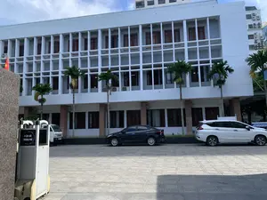 Bức ảnh chụp một tòa nhà văn phòng lớn, nhiều tầng với mặt tiền màu trắng có kết cấu dạng lưới bê tông. Phía trước tòa nhà là một khoảng sân được lát gạch, nơi có một vài chiếc ô tô đang đỗ, bao gồm một chiếc sedan màu đen, một chiếc SUV màu trắng và một chiếc xe tải nhỏ ở phía xa. Dọc theo sân là hàng cây cọ được trồng để trang trí. Ở phía bên trái của ảnh, có một thiết bị có bánh xe với biển hiệu ghi rõ chữ 'ĐIỆN LỰC THÀNH PHỐ'. Cũng ở phía bên trái, có thể thấy một phần của lá cờ đỏ sao vàng của Việt Nam. Khung cảnh cho thấy đây là một ngày trời trong, có mây. Không có người xuất hiện trong ảnh, chỉ có các phương tiện đang đỗ, cho thấy đây có thể là bãi đỗ xe của một cơ quan vào ban ngày.。（日本語訳）。写真には、コンクリートの格子状の構造を持つ白いファサードの、大きな多層階のオフィスビルが写っている。ビルの前にはタイル張りの広場があり、そこには黒いセダン、白いSUV、そして遠くに小型トラックを含む数台の車が駐車している。広場に沿って、装飾のためにヤシの木が植えられている。写真の左側には、「ĐIỆN LỰC THÀNH PHỐ」（市電力）という文字がはっきりと書かれた看板が付いた、車輪付きの装置がある。風景は、雲のある晴れた日であることを示している。写真には人は写っておらず、駐車している車両だけがあり、これは日中のどこかの機関の駐車場である可能性を示唆している。