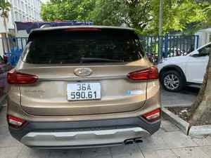 Hình ảnh chụp phía sau của một chiếc ô tô Hyundai Santa Fe màu vàng đồng đang đỗ trên vỉa hè hoặc bãi đậu xe lát gạch. Trên đuôi xe có các dòng chữ "SANTA FE", "2.2D" và một nhãn dán có logo Hyundai cùng dòng chữ "HYUNDAI THANH HÓA". Bên cạnh chiếc xe này là một phần của một chiếc ô tô khác màu trắng. Phía sau là cây xanh, một hàng rào sắt màu xanh và một tòa nhà có kiến trúc dạng lưới.。（日本語訳）。ブロンズゴールド色のヒュンダイ・サンタフェの後ろ姿を撮影した画像。車は歩道またはレンガ敷きの駐車場に停車している。車のリアには「SANTA FE」、「2.2D」の文字があり、そしてヒュンダイのロゴと「HYUNDAI THANH HOA」の文字が入ったステッカーが貼られている。後方には緑の木々、緑色の鉄柵、そして格子状の建築様式の建物がある。