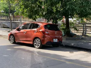 Hình ảnh chụp một chiếc xe ô tô hiệu Honda Brio RS màu cam đang đỗ bên lề một con đường nhựa. Chiếc xe được đỗ dưới bóng râm của một cây xanh lớn. Cạnh đó là vỉa hè và phía sau là một cây cầu có lan can bằng kim loại, bắc qua một con kênh hoặc sông. Ở phía xa, bên kia cầu, có một tòa nhà với tấm biển hiệu màu đỏ, trên đó có dòng chữ màu trắng có thể đọc được một phần: 'HỌC SINH KHÔNG ĐƯỢC RA SÁT MÉP...'. Trên đuôi xe ô tô có ghi chữ 'BRIO' và 'RS'.。（日本語訳）。オレンジ色のホンダ・ブリオRSが、アスファルトの道路の脇に駐車している写真。車は大きな緑の木の木陰に停められている。その隣は歩道で、後ろには金属製の手すりがついた橋が運河か川にかかっている。遠く、橋の向こうには赤い看板を掲げた建物があり、その看板には「HỌC SINH KHÔNG ĐƯỢC RA SÁT MÉP...」と部分的に読み取れる白い文字が書かれている。車の後部には「BRIO」と「RS」の文字が記されている。