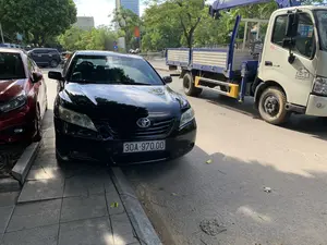 Bức ảnh chụp một cảnh đường phố vào ban ngày, có thể là ở thành phố Thanh Hóa. Ở vị trí trung tâm, một chiếc ô tô Toyota Camry màu đen đang đỗ một phần trên vỉa hè lát gạch. Bên cạnh và trên đường là một chiếc xe tải cẩu màu trắng, hiệu Hino, có thùng xe màu xanh và trắng. Trên cửa của chiếc xe tải này có các dòng chữ rõ ràng: "CÔNG TY ĐIỆN LỰC THANH HÓA", địa chỉ "96 TRIỆU QUỐC ĐẠT-P.ĐIỆN BIÊN-TP.TH", số điện thoại "ĐT: 0936 313 717", và các thông số kỹ thuật "4605KG", "2700KG", "7500KG", "3N". Ngoài ra còn có logo "HINO" và "EURO 3". Phía trước chiếc xe Camry là một phần của một chiếc ô tô khác màu đỏ. Khung cảnh xung quanh có nhiều cây xanh tươi tốt, và ở phía xa có thể thấy các tòa nhà, trong đó có một tòa nhà cao tầng. Có một vài người và phương tiện khác ở phía xa nhưng không rõ hoạt động cụ thể.。（日本語訳）。日中の街の風景を写した写真で、おそらくタインホア市での撮影です。中央には、黒いトヨタ・カムリがタイル張りの歩道に一部乗り上げて駐車しています。その隣の路上には、日野製の白いクレーン付きトラックがあり、荷台は青と白です。このトラックのドアには、「CÔNG TY ĐIỆN LỰC THANH HÓA」、住所「96 TRIỆU QUỐC ĐẠT-P.ĐIỆN BIÊN-TP.TH」、電話番号「ĐT: 0936 313 717」、そして技術仕様「4605KG」、「2700KG」、「7500KG」、「3N」という文字がはっきりと書かれています。その他に、「HINO」と「EURO 3」のロゴもあります。カムリの前方には、別の赤い車の一部が見えます。周囲の風景には青々とした緑の木々が多く、遠くには高層ビルを含む建物が見えます。遠くには数人の人物や他の車両がいますが、具体的な活動ははっきりしません。