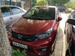 Bối cảnh là một khu vực đỗ xe trên vỉa hè hoặc trước một tòa nhà trong một khu đô thị. Trọng tâm của hình ảnh là một chiếc ô tô Honda màu đỏ, phiên bản RS, đang đỗ. Chiếc xe này đỗ gần một thân cây được trang trí bằng dây đèn LED. Xung quanh có các phương tiện khác, bao gồm một chiếc ô tô màu bạc ở bên trái và một chiếc ô tô màu đen ở bên phải. Ở phía sau, có thể nhìn thấy một phần của một tòa nhà hiện đại và quang cảnh đường phố với nhiều xe cộ hơn, trong đó có một người mặc đồng phục màu cam đang đứng cạnh một chiếc xe máy màu vàng.。（日本語訳）。背景は、都市部にある歩道上または建物の前の駐車エリアです。画像の中心は、駐車している一台の赤いホンダ車、RSバージョンです。この車は、LEDライトの dây で飾られた木の幹の近くに駐車しています。周りには他の車両があり、左側には一台の銀色の車、右側には一台の黒い車が含まれます。後方には、近代的な建物の一部と、さらに多くの車両がある街の風景が見え、その中にはオレンジ色の制服を着た人が黄色いバイクの隣に立っています。