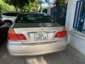 Bức ảnh chụp phía sau của một chiếc ô tô Toyota Camry 2.4G màu bạc đang đỗ trên vỉa hè lát gạch. Trên thân xe có các ký hiệu '2.4G' và 'CAMRY' cùng với logo của Toyota. Chiếc xe đỗ cạnh một hàng rào sắt màu xanh và một bức tường trắng. Xung quanh có nhiều cây xanh và có thể thấy một phần của các chiếc xe khác đang đỗ gần đó. Một chiếc lá vàng nằm trên cốp xe.。（日本語訳）。タイル張りの歩道に駐車している、銀色のトヨタ・カムリ2.4Gの後ろ姿の写真。車体には「2.4G」と「CAMRY」の記号とトヨタのロゴがある。車は青い鉄柵と白い壁の隣に駐車している。一枚の黄色い葉が車のトランクの上にある。