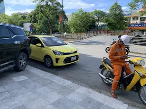 Bức ảnh chụp một khung cảnh đường phố vào ban ngày, có thể là tại thành phố Thanh Hóa, dựa trên bối cảnh. Ở tiền cảnh, một người đàn ông mặc bộ đồ bảo hộ lao động màu cam, đội mũ bảo hiểm trắng, đang ngồi trên chiếc xe máy Honda màu vàng và sử dụng điện thoại di động. Bên cạnh vỉa hè, một chiếc ô tô SUV màu đen và một chiếc sedan Kia màu vàng đang đỗ. Ở hậu cảnh, có một cây cầu nhỏ dành cho người đi bộ với lan can màu trắng. Phía sau cây cầu là một tòa nhà lớn, là một trường học. Trên cổng và tòa nhà có nhiều biển hiệu và biểu ngữ. Một biểu ngữ lớn có thể đọc được một phần: 'TRƯỜNG TRUNG HỌC CƠ SỞ' và dòng chữ bên dưới 'ĐẢNG BỘ, CHÍNH QUYỀN VÀ NHÂN DÂN PHƯỜNG ĐIỆN BIÊN QUYẾT TÂM TRỞ THÀNH “ĐƠN VỊ ANH HÙNG LAO ĐỘNG TRONG THỜI KỲ...”'. Ngoài ra còn có một biển báo giao thông cấm dừng và đỗ xe với ký hiệu '20M'. Khung cảnh có nhiều cây xanh và một tòa nhà cao tầng hiện đại ở phía xa.。（日本語訳）。背景から、おそらくタインホア市の、昼間の街の風景を写した写真です。前景には、オレンジ色の作業着を着て白いヘルメットをかぶった男性が、黄色のホンダ製バイクに座って携帯電話を使用しています。歩道の横には、黒いSUVと黄色の起亜製セダンが駐車しています。背景には、白い手すりのある小さな歩道橋があります。橋の後ろには大きな建物があり、それは学校です。門と建物には多くの看板や横断幕があります。大きな横断幕の一部が読み取れます：「中学校」、そしてその下の行には「ディエンビエン区の党委員会、政府、人民は『…期の労働英雄単位』になることを決意する」。また、「20M」という記号の付いた駐停車禁止の交通標識もあります。風景には多くの緑の木々と、遠くには近代的な高層ビルがあります。